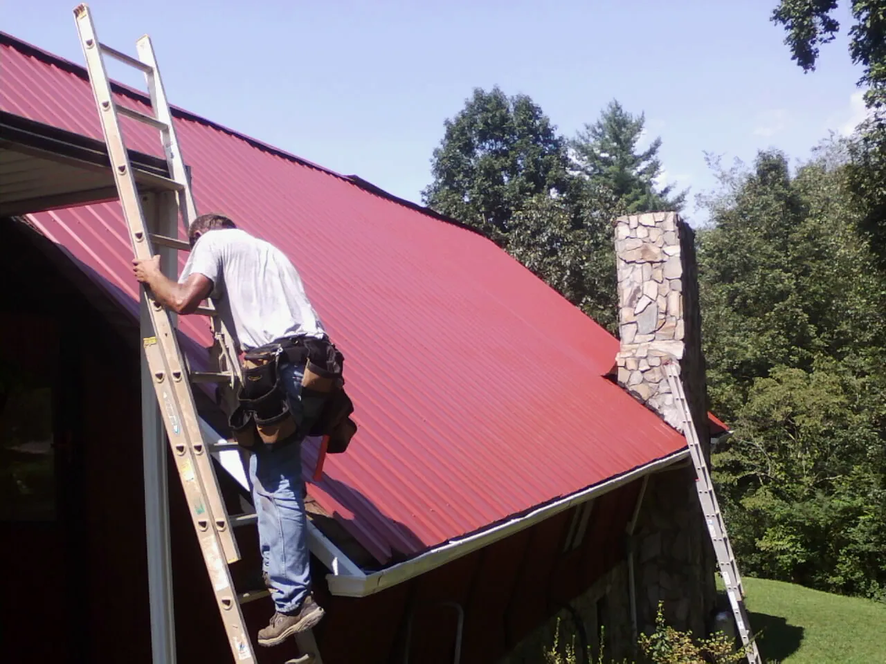 Roofer on a ladder beside a red metal roof and stone chimney — Soffit & Fascia Repair by Roofing Brothers in Red Boiling Springs